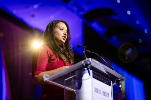 Speaker in red dress at podium during ACE Scholarships Women's Luncheon Denver Colorado